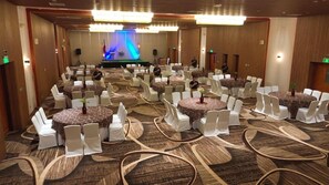 Un salón de conferencias con varias mesas redondas, sillas blancas y un escenario con fondo azul.
