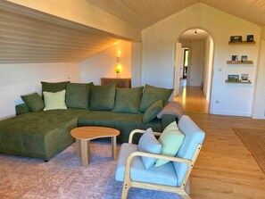 A living room with a sectional sofa, a small round table, and a wooden chair. There is a lamp on a side table, and a hallway leading to another room. - Alpenblick.Hoch3 Vacation rental with two separate bedrooms and three balconies facing east-southwest (Aschau im Chiemgau)