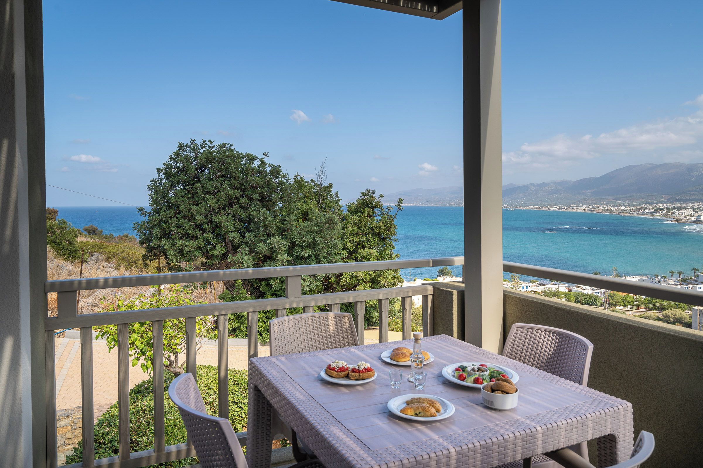 A balcony with a table set for dining, overlooking a coastal view with a clear sky and distant mountains.