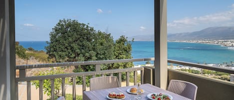 A balcony with a table set for dining, overlooking a coastal view with a clear sky and distant mountains.