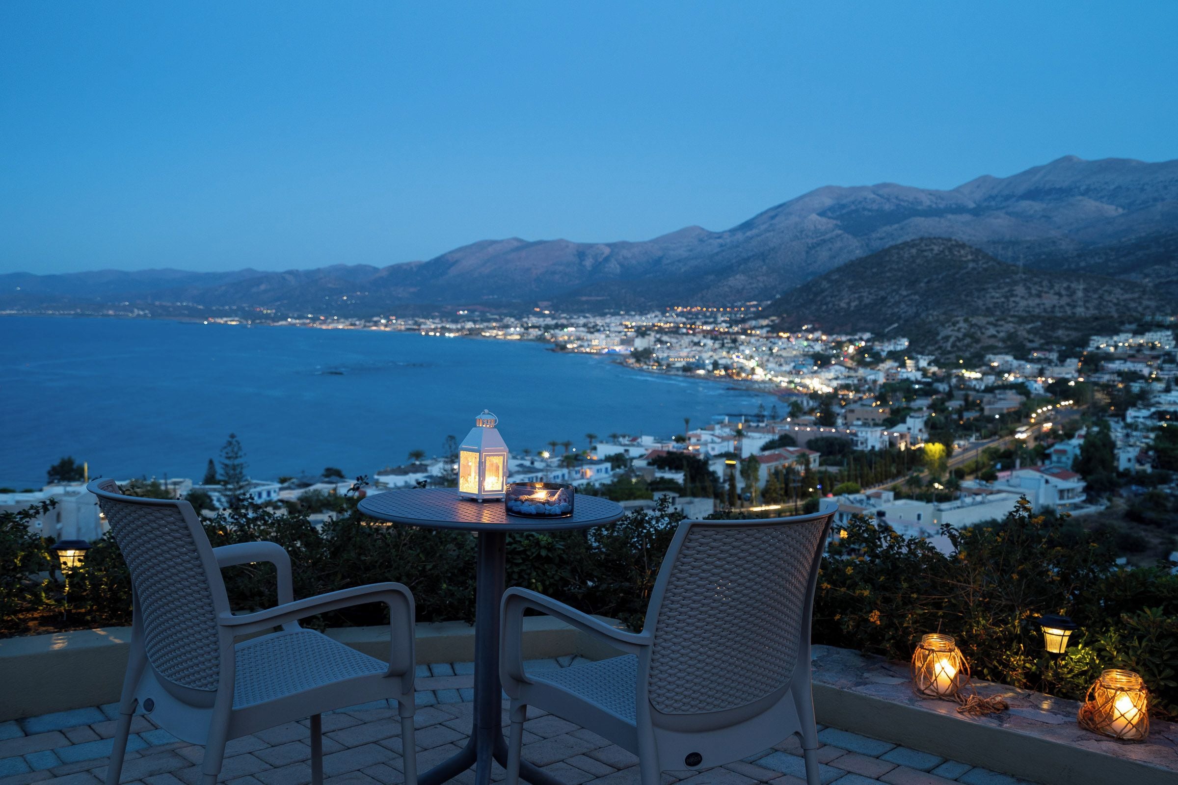 Una terraza frente al mar con dos sillas, una mesa y vistas a un pueblo costero al anochecer.