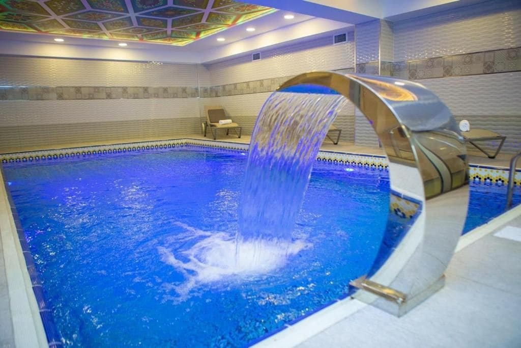 Ein Schwimmbecken mit Wasserfall, umgeben von Fliesenwänden und einer Decke mit geometrischem Muster.