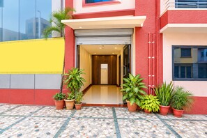 Entrada de hotel con piso de baldosas, plantas en macetas y fachada roja y amarilla.