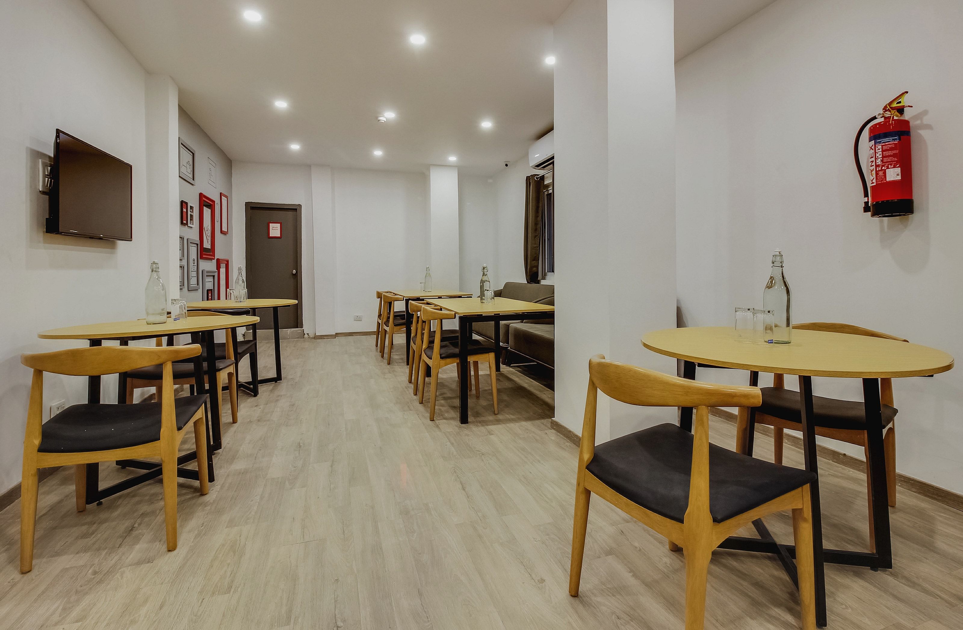 A hotel dining area with multiple tables and chairs, a fire extinguisher mounted on the wall, and a door with a sign.