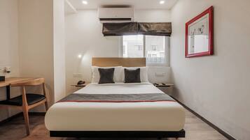 A hotel room with a bed, bedside table, a desk, and an air conditioning unit.