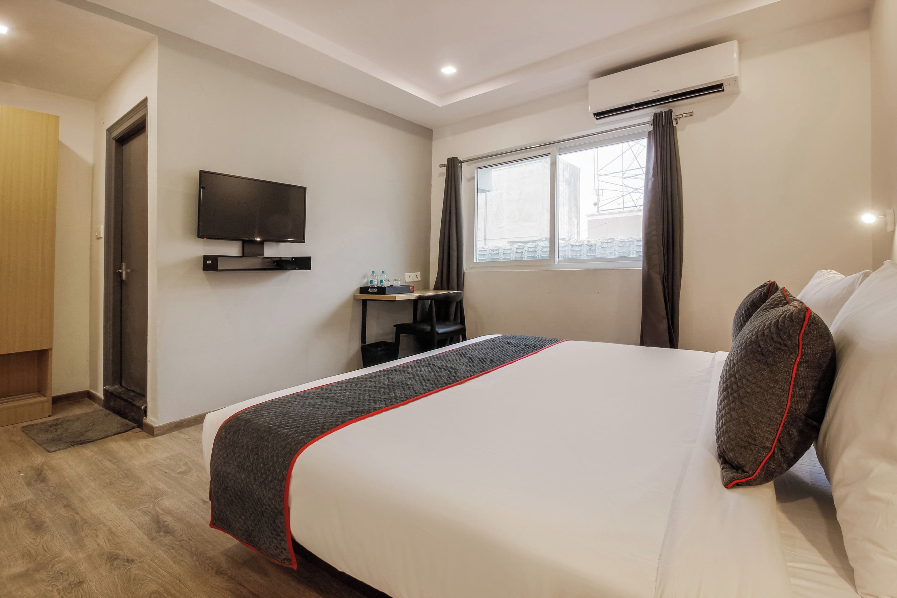 A hotel room with a bed, a television mounted on the wall, a window with curtains, and an air conditioning unit.