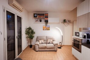 Ein gemütlicher Wohnbereich mit Küchenzeile, einer Couch mit Zierkissen, einer Topfpflanze und einem an der Wand befestigten Regal mit Büchern und Dekoration.