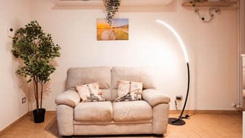 A neutral-toned sofa with two decorative pillows, a potted plant, a wall-mounted picture, and a floor lamp in a room with wooden flooring.