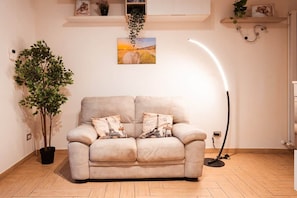 A neutral-toned sofa with two decorative pillows, a potted plant, a wall-mounted picture, and a floor lamp in a room with wooden flooring.