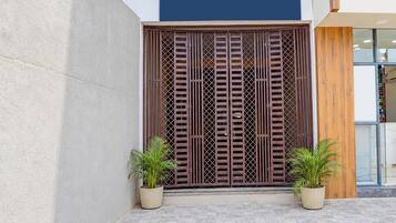 Una entrada de edificio con un portón metálico grande, dos plantas en macetas y un pavimento de baldosas.