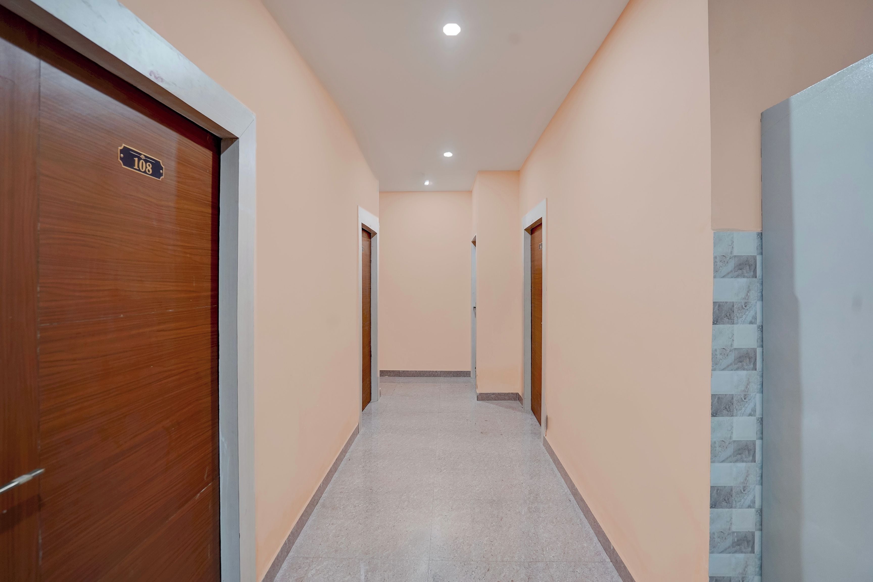 A corridor with a wooden door on the left, a tiled wall on the right, and a tiled floor.