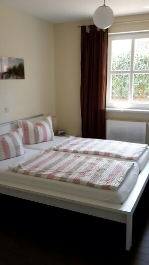 A bed with a white frame and pink and white striped bedding, a bedside table with a clock, and a window with brown curtains.