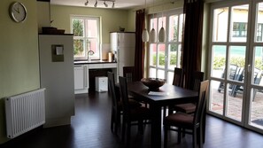 A dining area with a table and chairs, a clock on the wall, and a view of the outdoors through large windows.