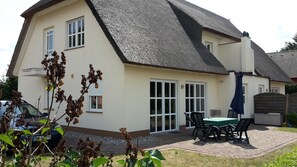 Une maison avec un toit à forte pente, une terrasse avec une table et des chaises, et une voiture garée dans l’allée.