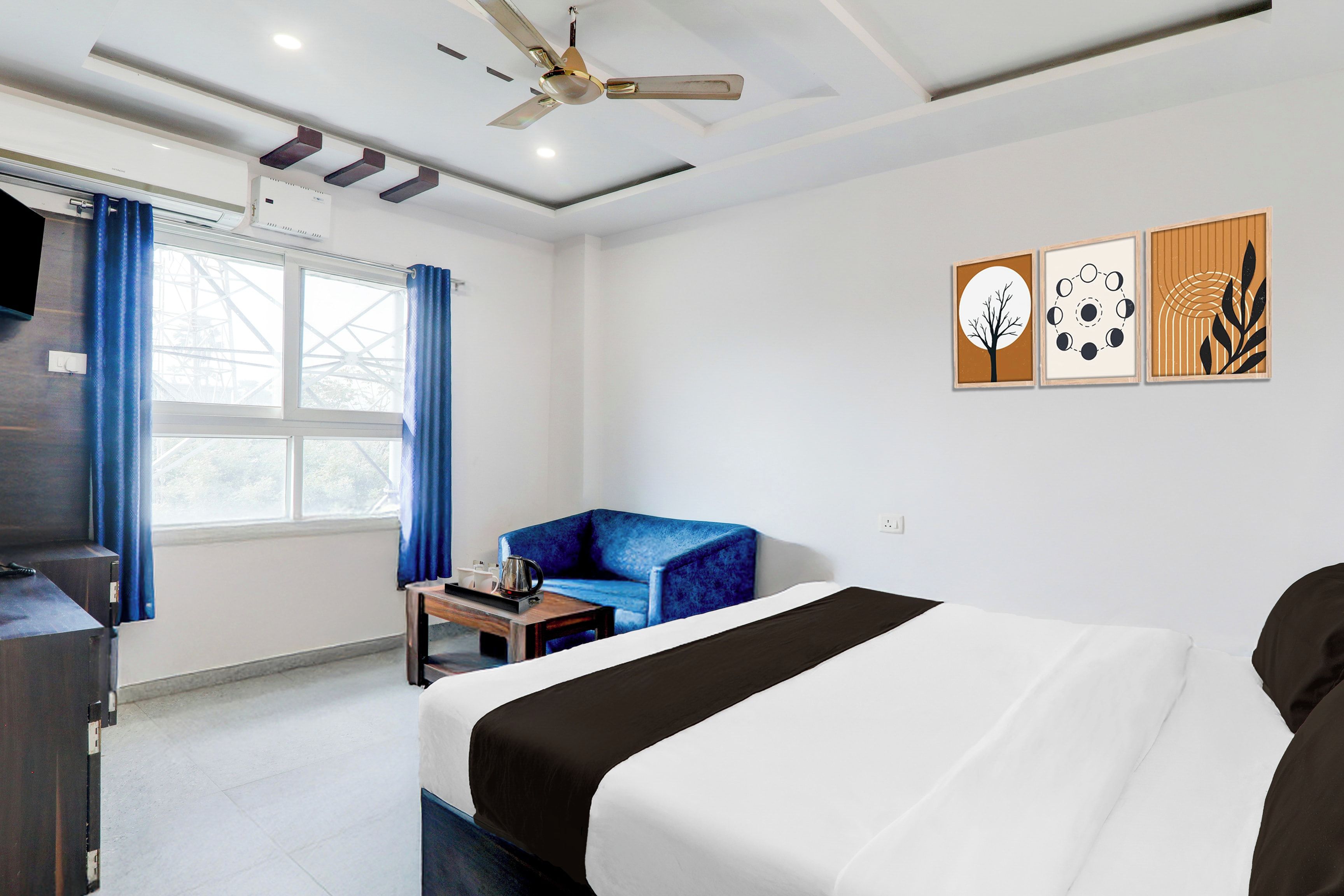 Habitación de hotel con cama, sofá azul, una mesita con bandeja, ventilador de techo y cuadros en la pared.