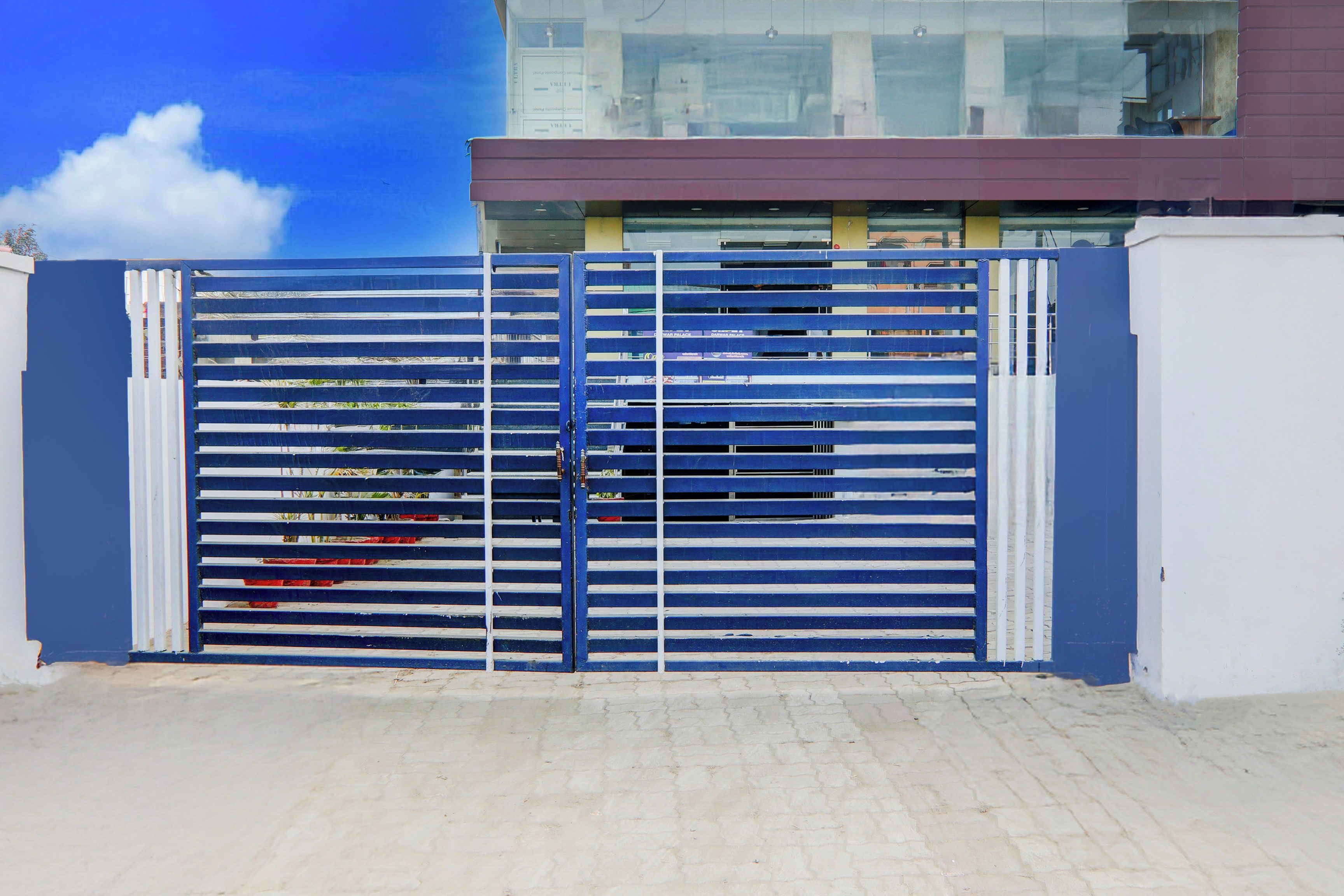 Una verja azul de metal con barras horizontales, suelo empedrado y un edificio con fachada de vidrio al fondo.