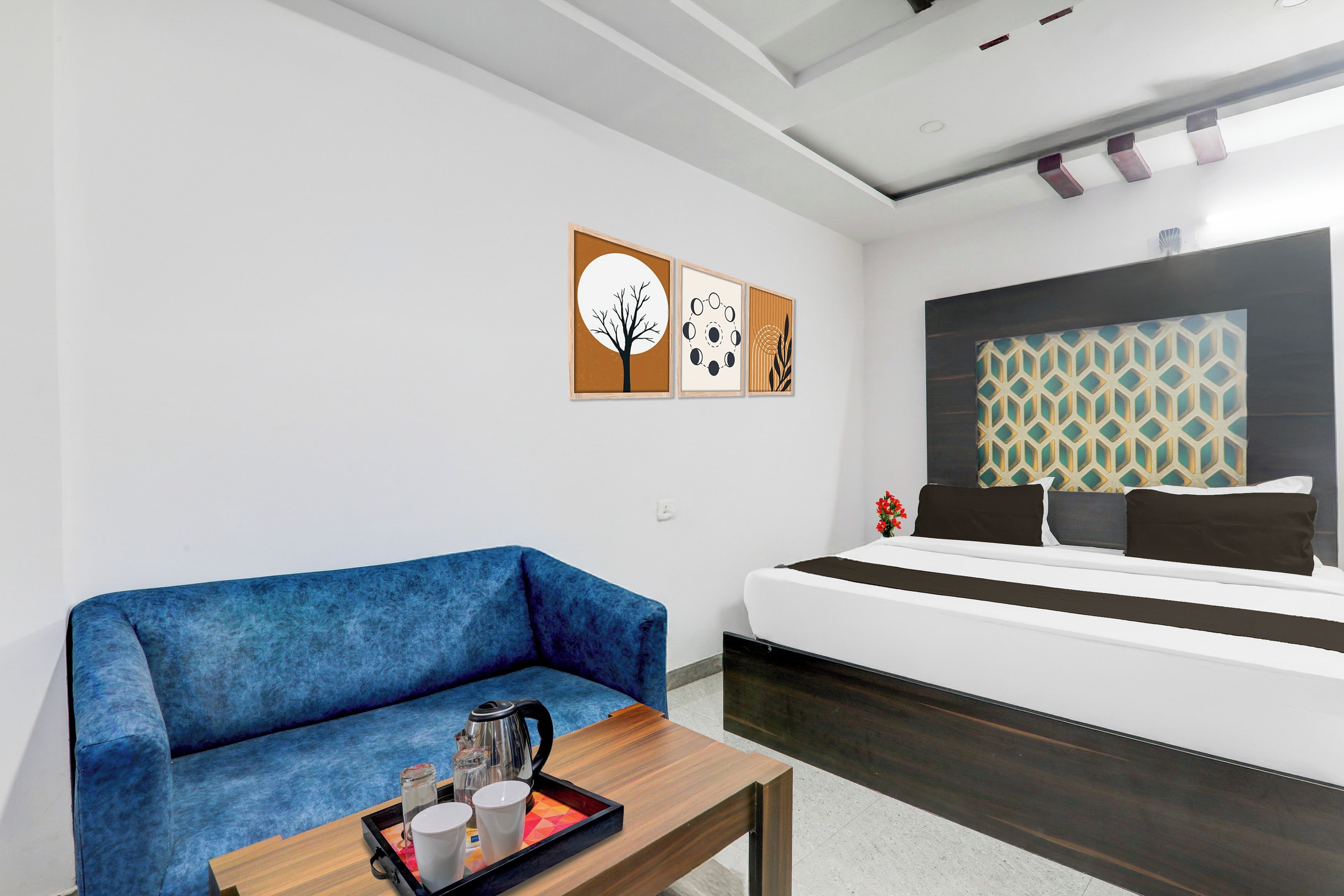 Quarto de hotel moderno com sofá azul, mesa de centro de madeira com bandeja de xícaras e chaleira, cama com cabeceira estampada e duas obras de arte nas paredes.