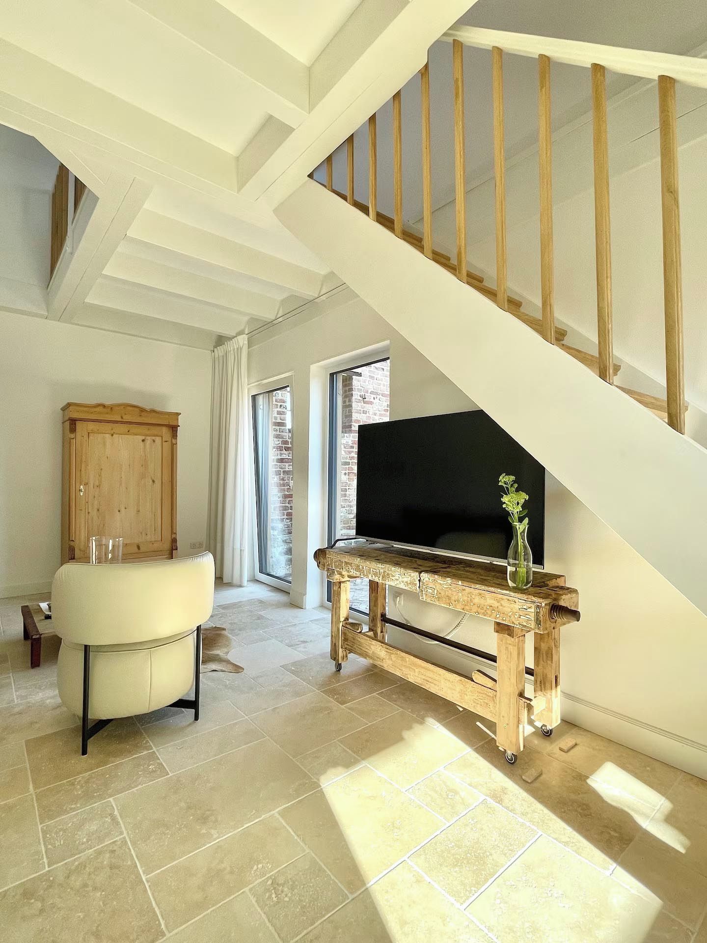 Une pièce spacieuse avec un plafond haut, un escalier en bois, un téléviseur posé sur une console en bois et un fauteuil en cuir.