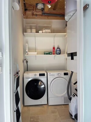 A laundry room with two front-loading washing machines, a shelf with cleaning supplies, and a hose for water connection. - Small country house surrounded by greenery, peace and quiet, nature, a break from it all, breakfast included (Fröndenberg/Ruhr)