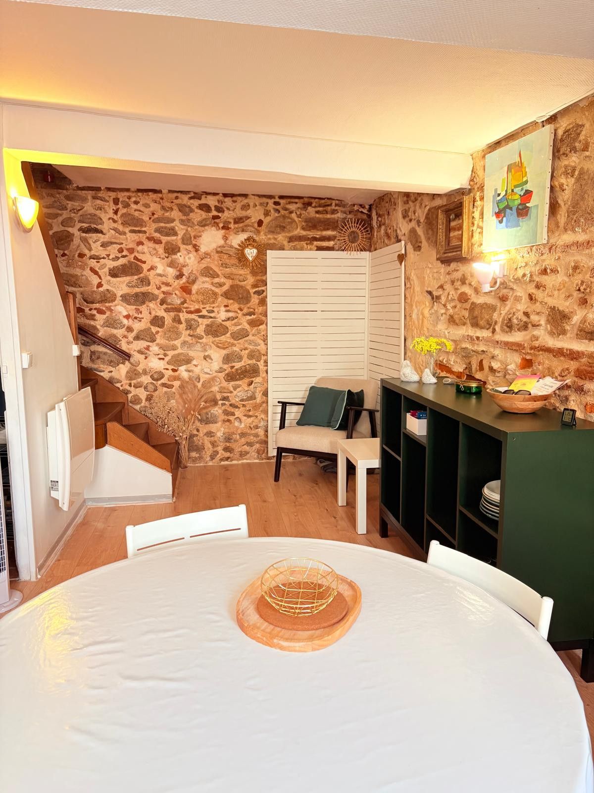 Une pièce chaleureuse avec un mur en pierre, une table blanche agrémentée d’un élément décoratif, une chaise et une petite étagère avec quelques objets.