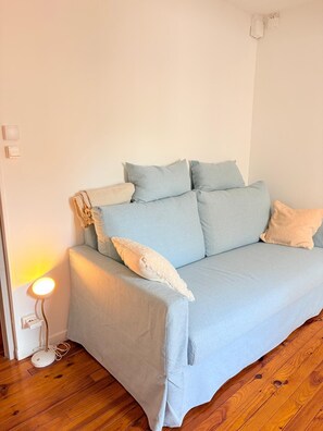 A light blue sofa with multiple cushions against a plain wall, a floor lamp with a white shade, and a wooden floor. - St Cyprien Cocon in the heart of the village (Saint-Cyprien)