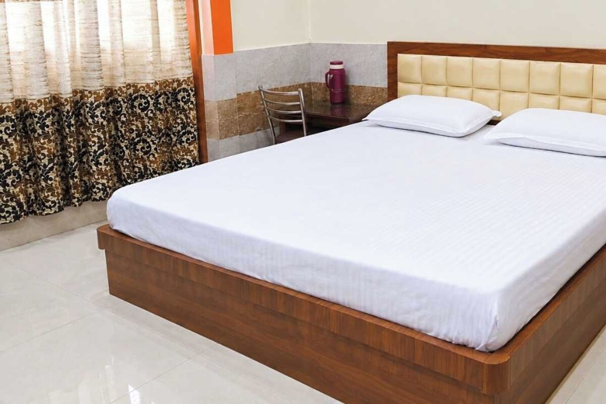 Een netjes opgemaakt bed met houten frame en witte beddengoed, een nachtkastje met een rode mok, en een stoel in een hotelkamer.