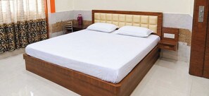 Een netjes opgemaakt bed met houten frame en witte beddengoed, een nachtkastje met een rode mok, en een stoel in een hotelkamer.