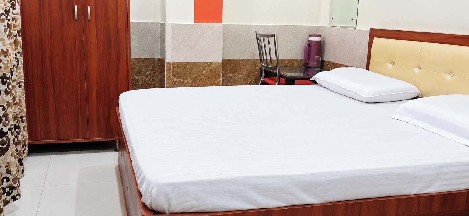 Un letto rifatto con lenzuola bianche e una testiera in legno, una sedia con un cuscino rosso e un armadio con specchio.