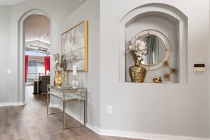 A hallway with a glass console table, a framed abstract painting, a mirror with a gold frame, and a vase with flowers. - Four Palms Pool Retreat | 4BR, Private Pool & Game (Kissimmee)