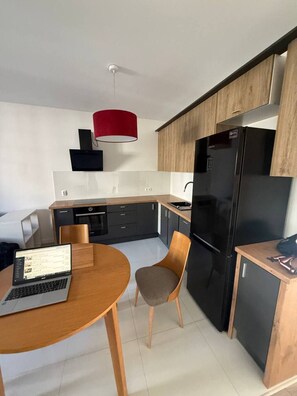 A modern kitchen with a black refrigerator, wooden cabinets, a red pendant light, and a laptop on a wooden table. - Lumina 2BR Apt w/ Parking & Balcony  (Warszawa)
