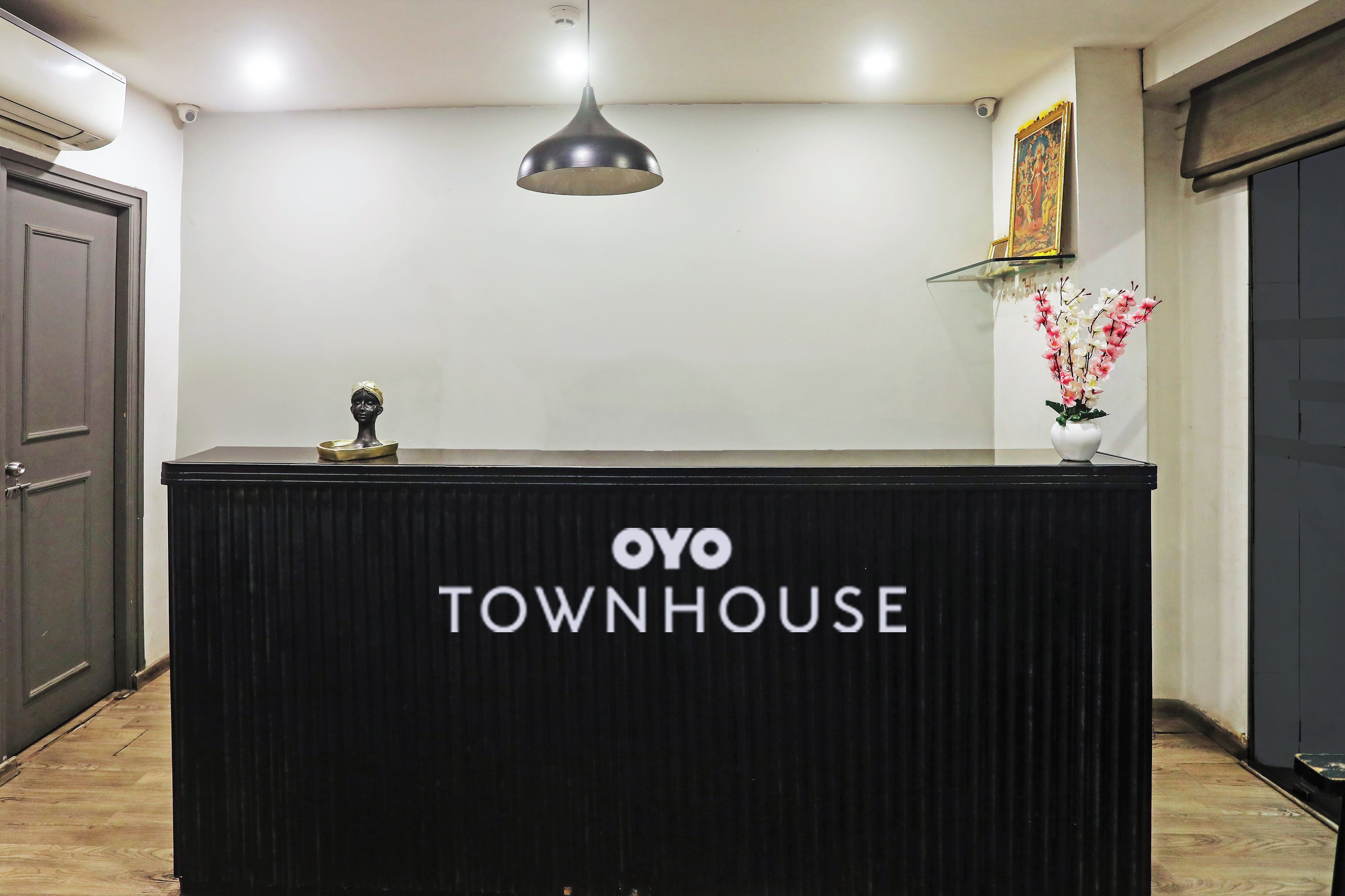 Una recepción con letrero 'OYO TOWNHOUSE', una lámpara colgante, un cuadro en la pared y un florero con flores.