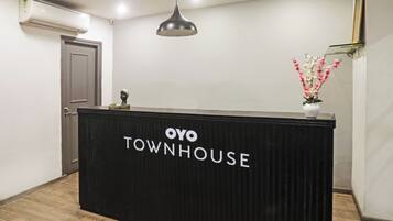Una recepción con letrero 'OYO TOWNHOUSE', un aire acondicionado instalado en el techo, una lámpara colgante y un jarrón con flores.