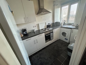 A compact kitchen with a stove, oven, washing machine, and a window with blinds. - Central London Studio Apartment- walking distance from the London Eye/Big Ben (London)