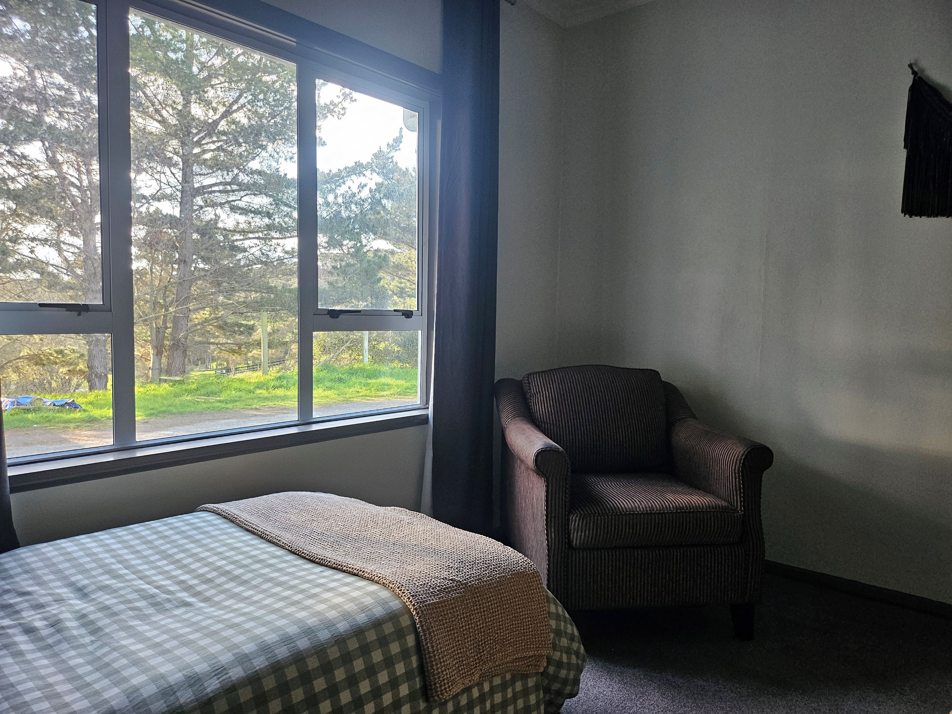 A room with a bed, a chair, and a window with a view of trees and a path.