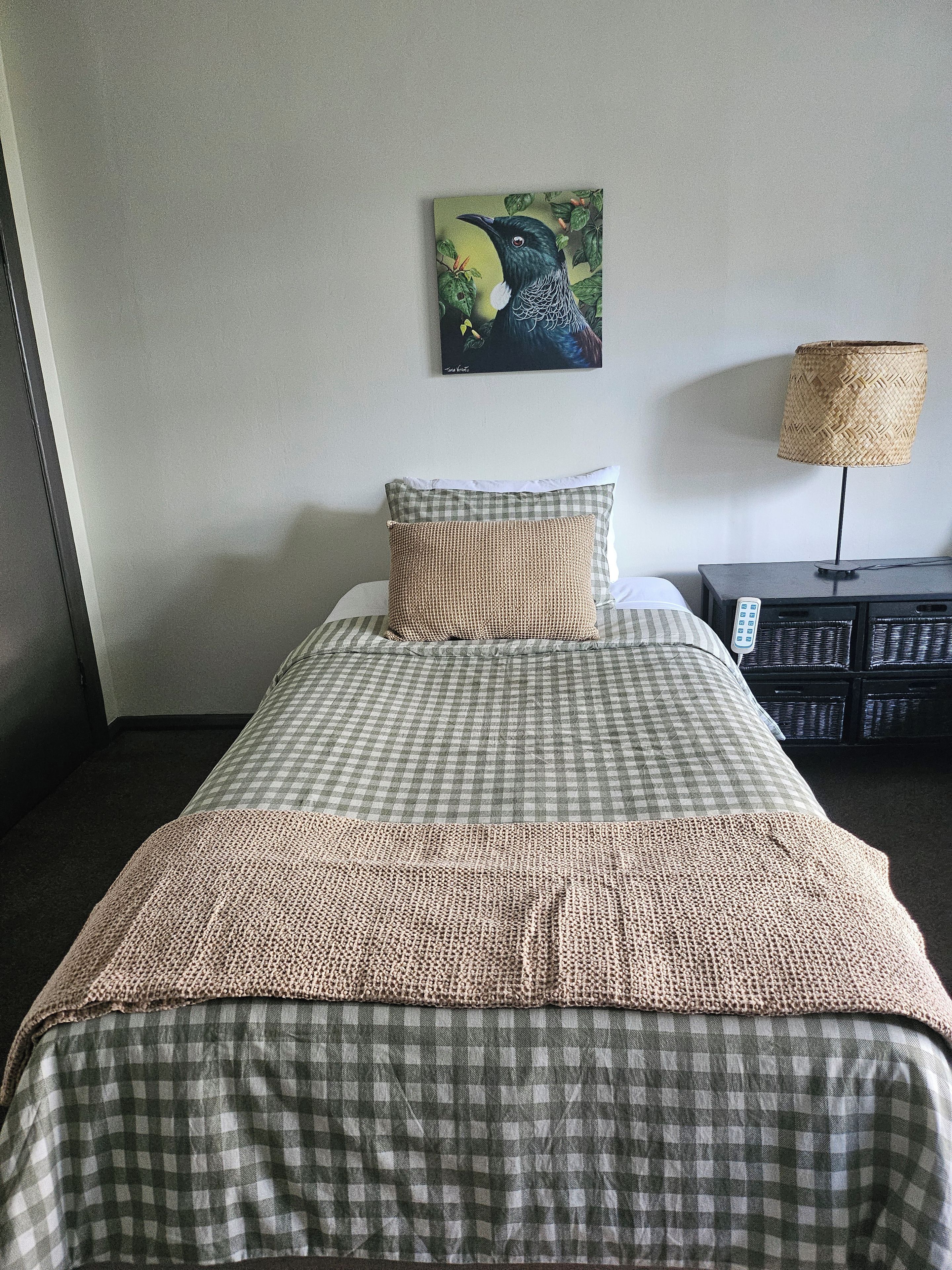 A hotel room with a bed, a checkered bedspread, a pillow, a wall-mounted painting, a lamp, and a side table with a remote control.