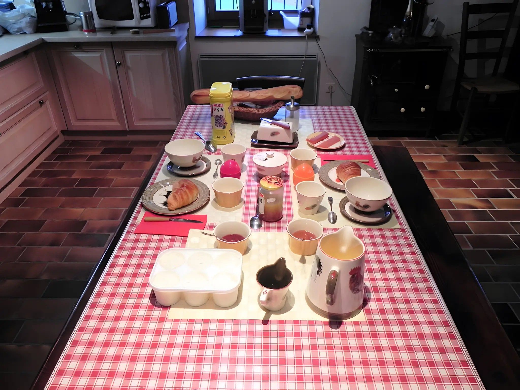 Een keuken met een rood-wit geruit tafelkleed, diverse keukenapparaten en een eettafel gedekt met borden, kommen en kopjes.