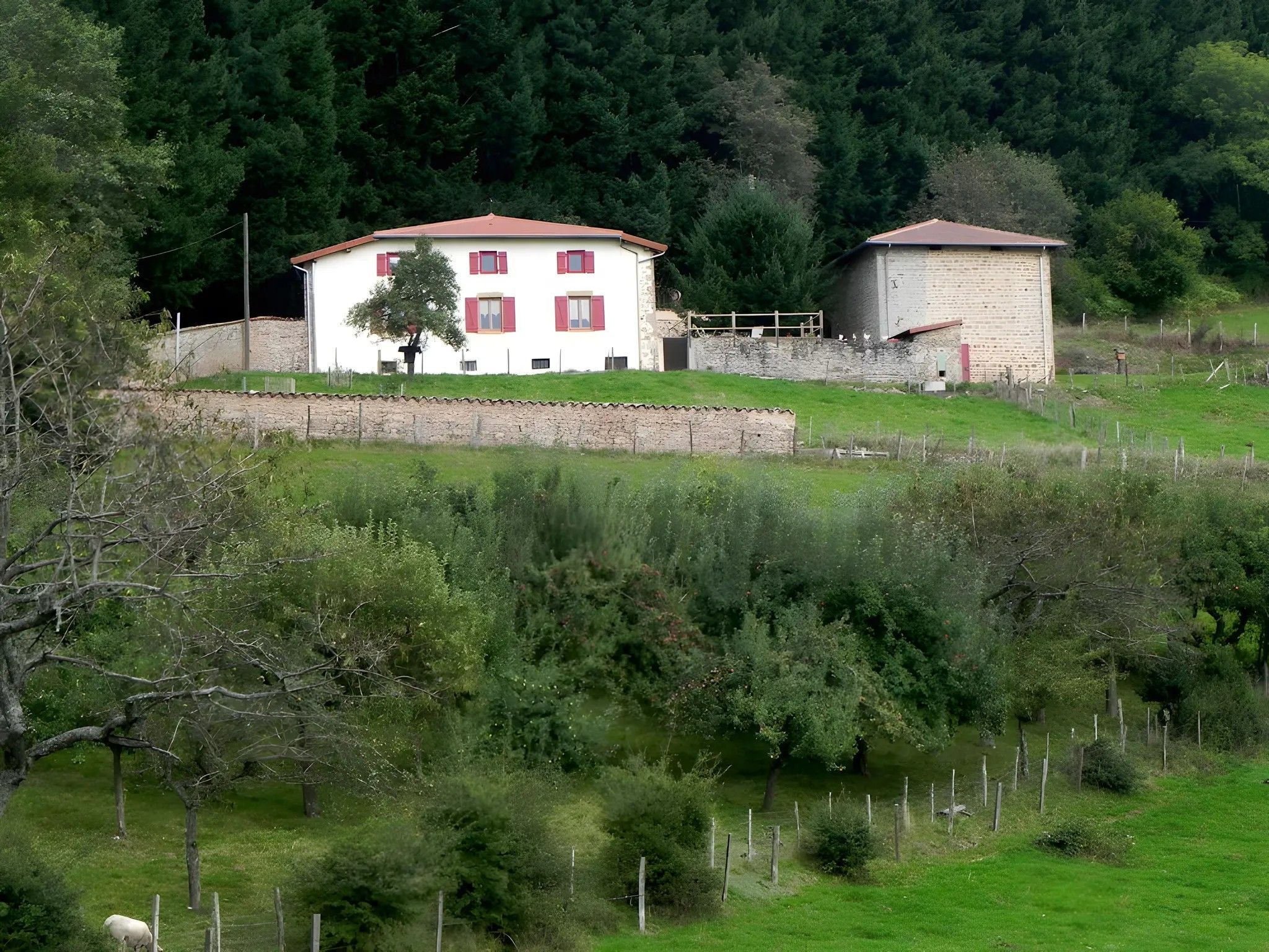 Un bâtiment à deux étages, avec un toit rouge et des murs blancs, entouré de verdure et d’un mur de pierre.
