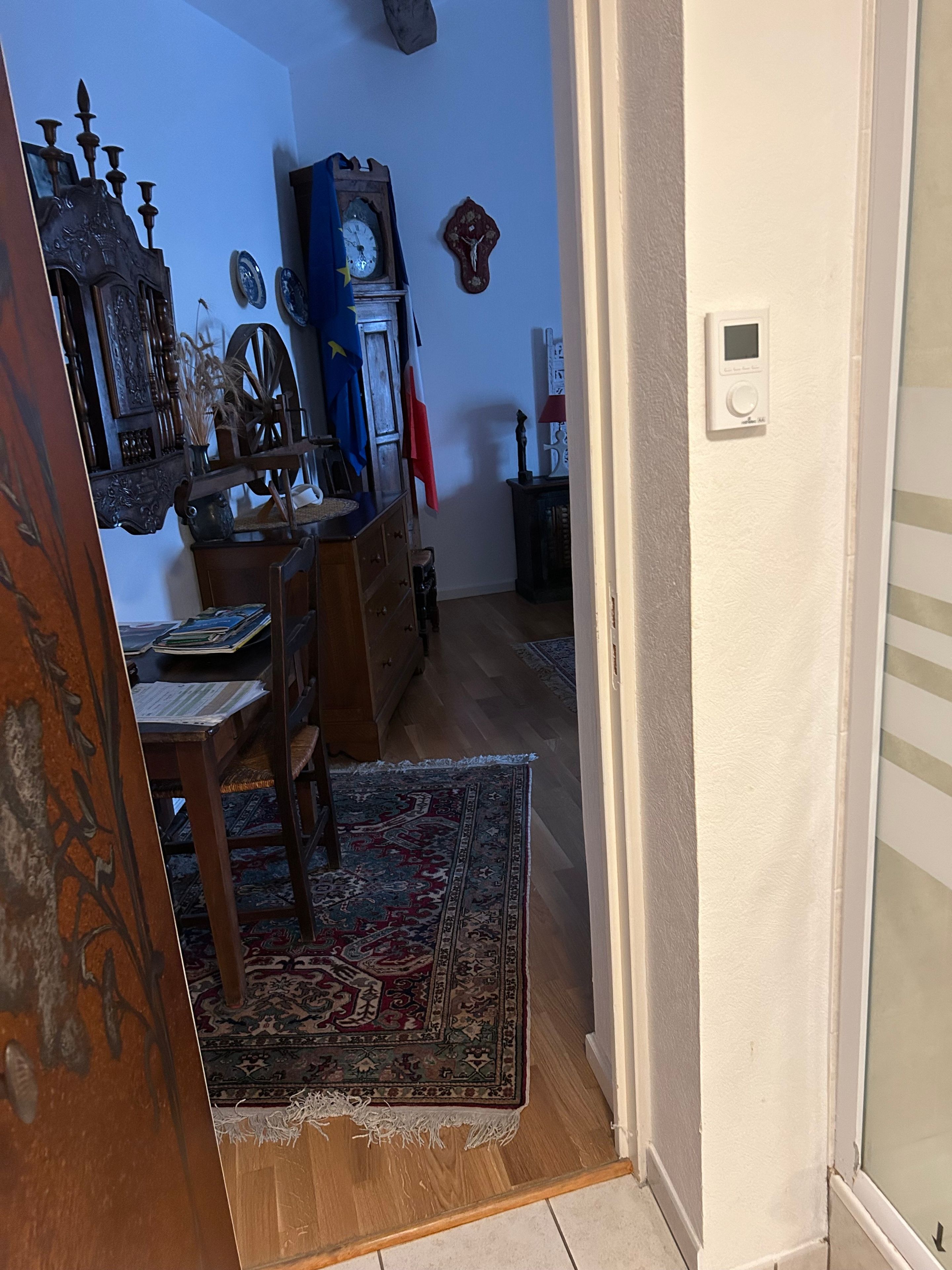 Een kamer met een houten vloer, een kamerbreed tapijt met dessin, een houten tafel met stoelen, een klok en een muurbevestigde thermostaat.