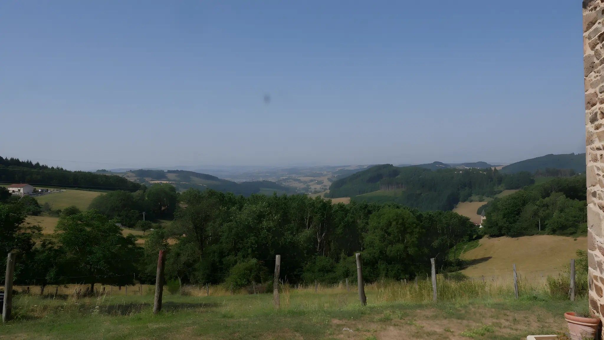A rural landscape with a stone wall on the right, a fenced area in the foreground, and a valley with scattered buildings in the distance.