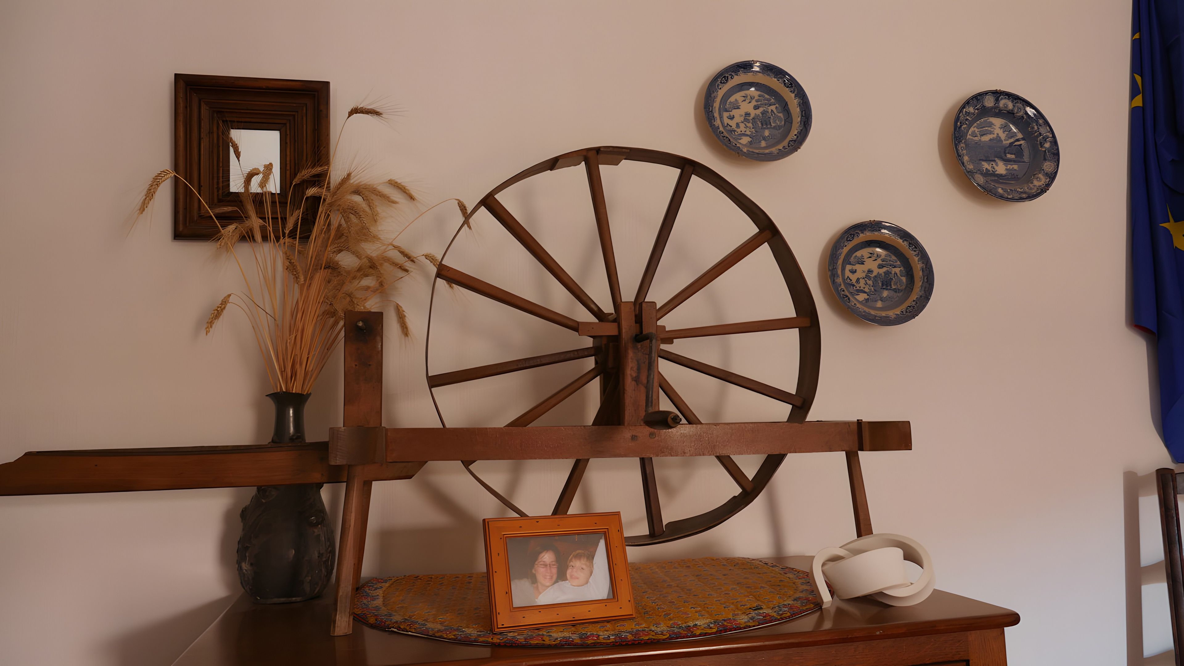 Una silla de madera rústica con una rueda grande, un florero con plantas secas y una foto en un marco sobre una mesa.