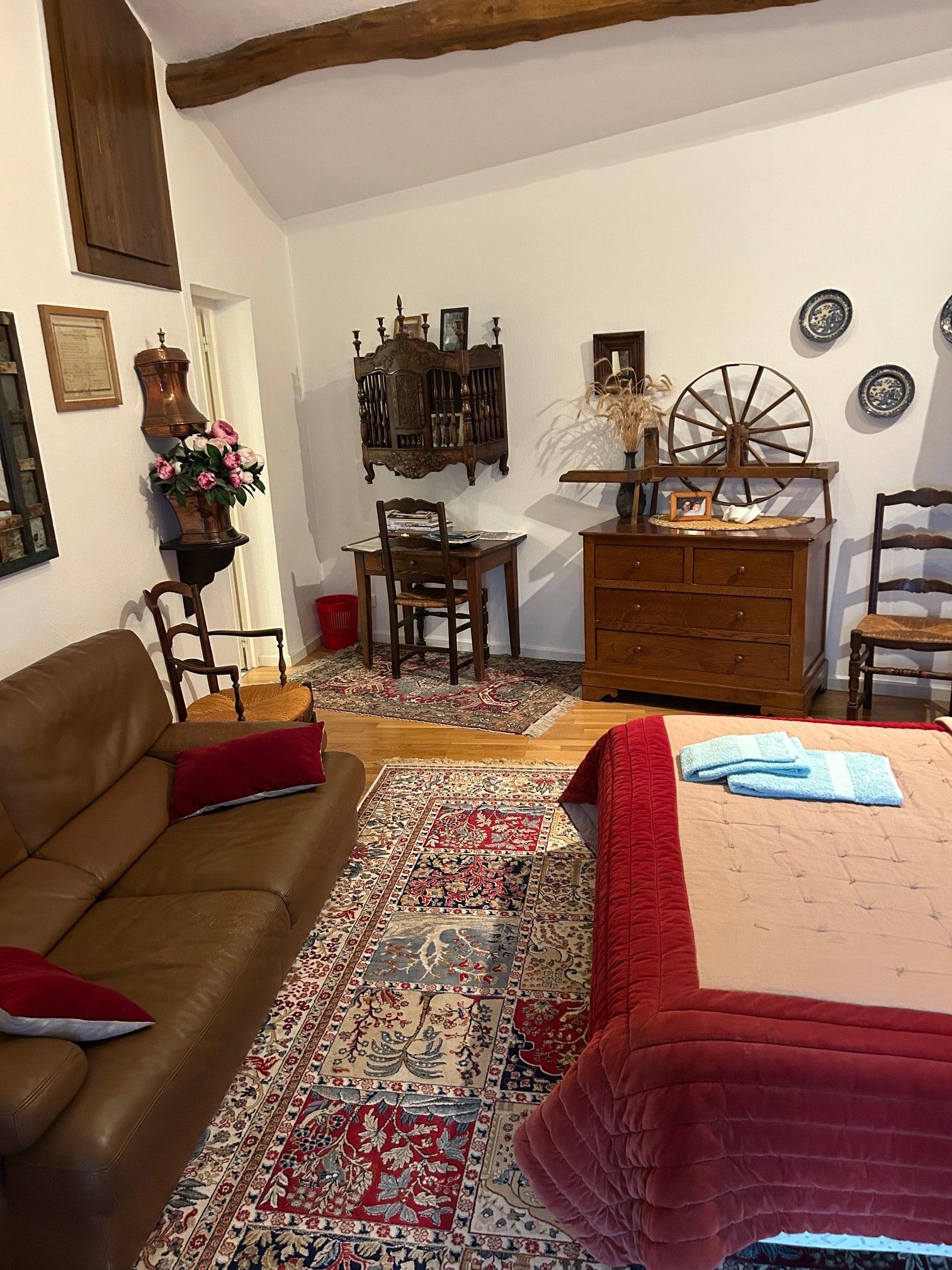 Una habitación tradicional con techo de madera, sofá de cuero marrón, cama con colcha acolchada roja, escritorio de madera y alfombra estampada.