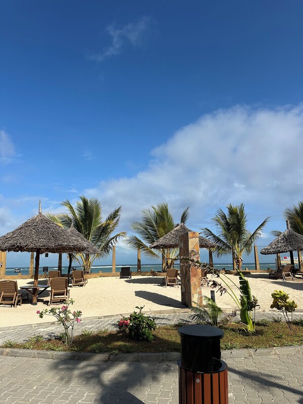 A beachside seating area with thatched umbrellas and lounge chairs, a trash bin, and a view of the ocean.