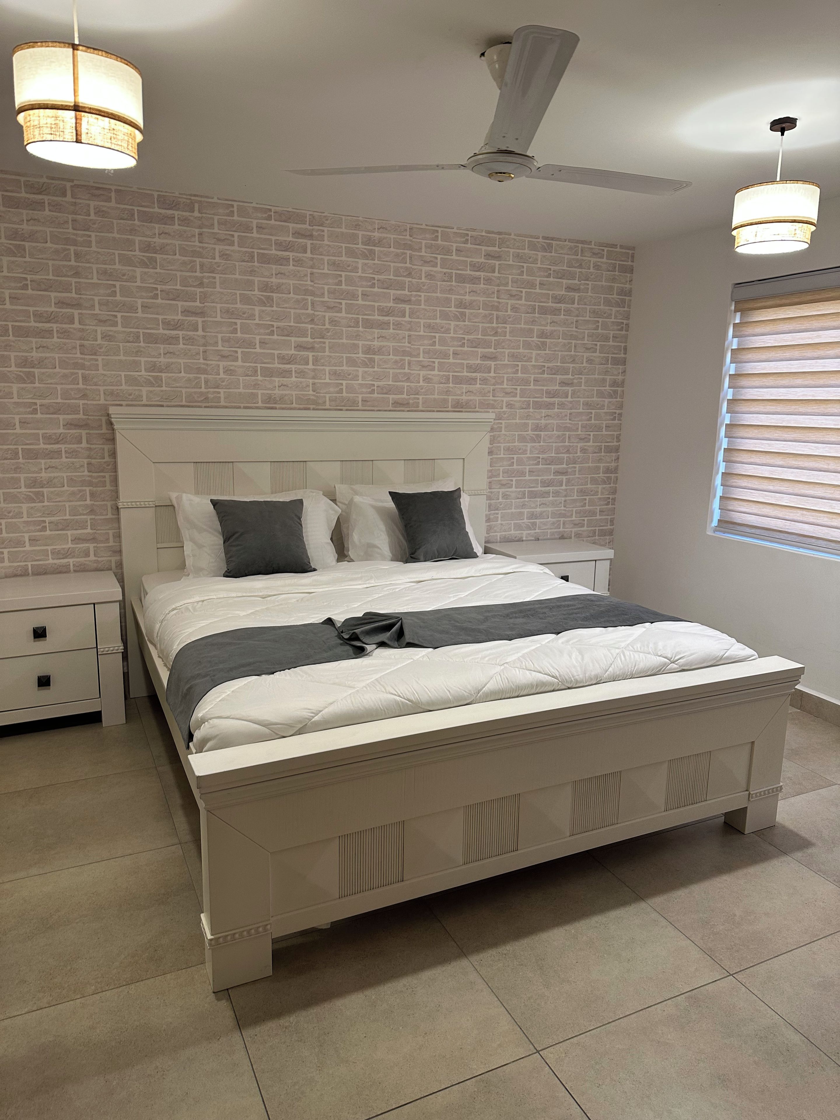 Un dormitorio con cama blanca, mesita de noche, ventilador de techo y pared con diseño de ladrillo.