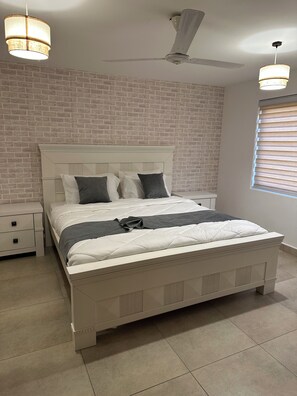 A bedroom with a white bed, a nightstand, a ceiling fan, and a brick-patterned wall.