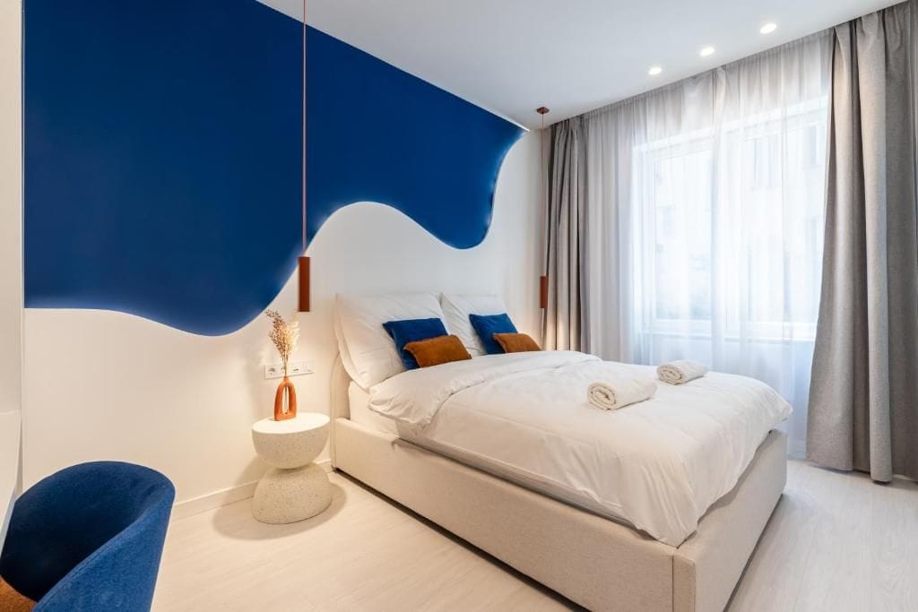 Ein modernes Schlafzimmer mit einem großen Bett, einer blauen Akzentwand, einem runden Beistelltisch und einem blauen Sessel.