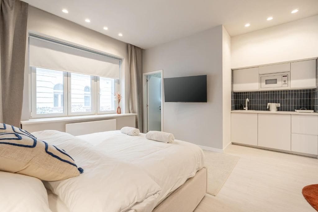 A modern bedroom with a large bed, a flat-screen TV mounted on the wall, and a kitchenette area with a microwave and cabinets.