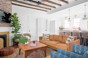 A modern living room with a sofa, coffee table, and chairs. There is a fireplace, a large plant, and a kitchen area visible in the background. - Silver Spring | The Crown Jewel Suite (Charleston)