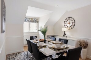 A dining area with a white table set for four, black chairs, a large wall clock, and a potted plant. - Charming 2-bedroom apartment in New Barnet perfect for relaxing stays (New Barnet)