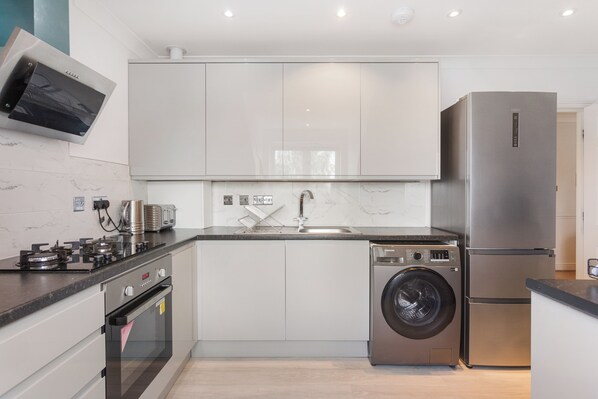 A modern kitchen with a built-in oven, a stainless steel refrigerator, a washing machine, and a flat-screen TV mounted above the sink. - Charming 2-bedroom apartment in New Barnet perfect for relaxing stays (New Barnet)