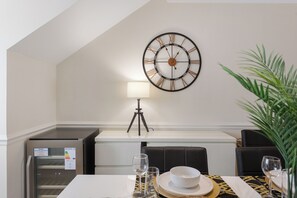 A dining area with a wall-mounted clock, a lamp, a small refrigerator, and a table set for two with plates, glasses, and a bowl. - Charming 2-bedroom apartment in New Barnet perfect for relaxing stays (New Barnet)